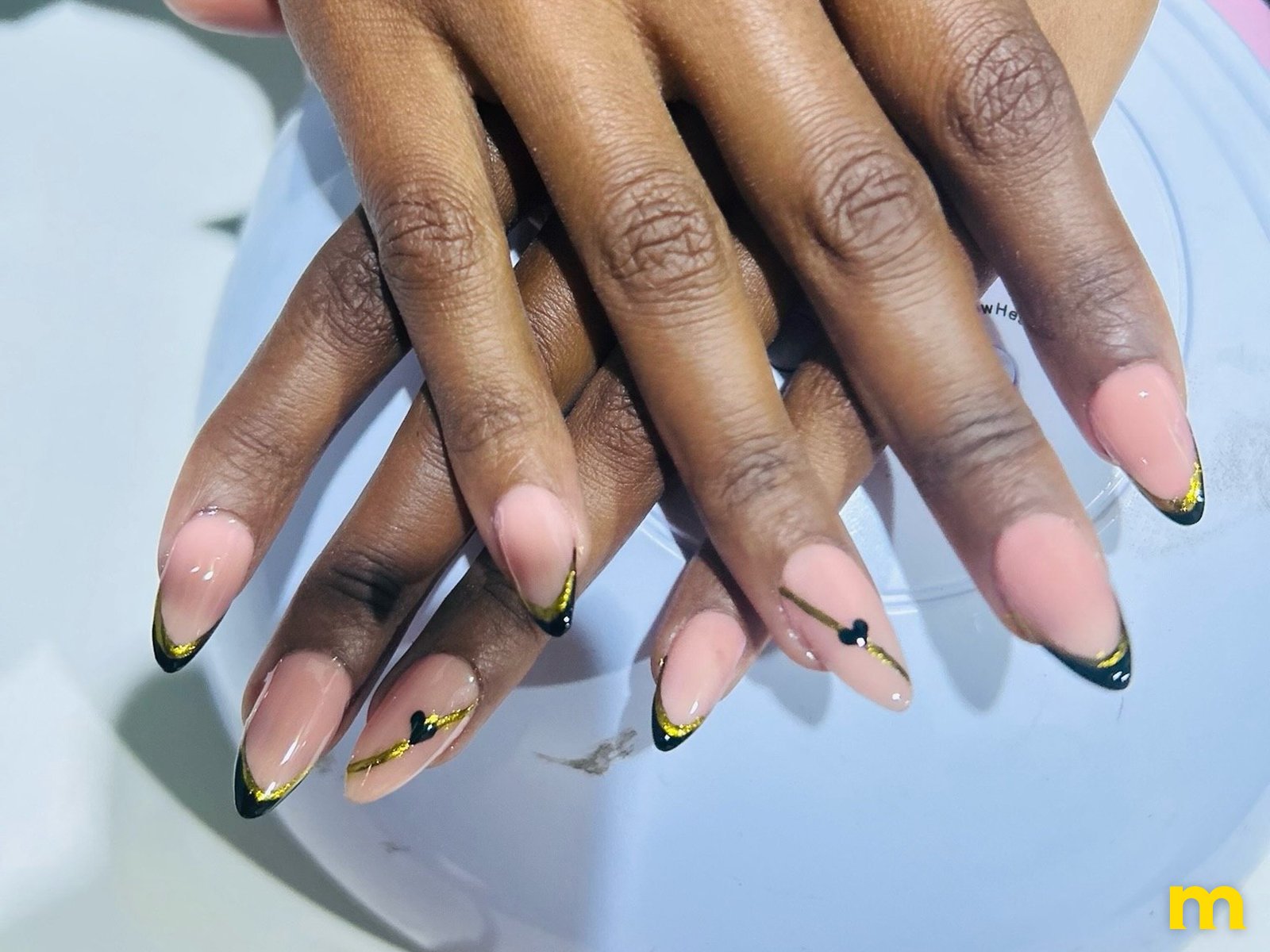 Unhas amendoadas nude com pontas pretas, linhas douradas e um coração preto, pintadas pelo Nélio